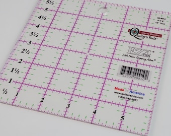 May include: A clear, square quilting ruler with a grid pattern in purple and green. The ruler has measurements in inches and features the "Quilter's Rule" logo and the text "Made in America". It is 8 inches by 8 inches.