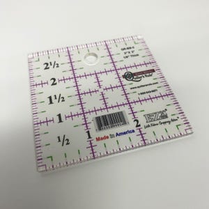 以下が含まれることがあります： 紫と緑の印が付いた透明なプラスチック製のキルト定規。定規は 7.6 cm &times; 7.6 cm で、厚さは 3 mm です。中央に穴があり、右下隅には「EZ2C with Fabric Cropping Action」というテキストが印刷されています。右上隅には「Quilter's Rule」というテキストが印刷されています。