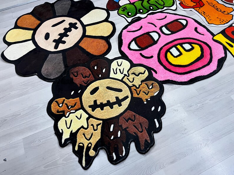 May include: Three colorful, cartoon-style rugs with different designs. The first rug is a black and brown flower with a yellow center and a stitched-up mouth. The second rug is a pink face with a yellow smile and red eyes. The third rug is a black and brown flower with a yellow center and a stitched-up mouth.
