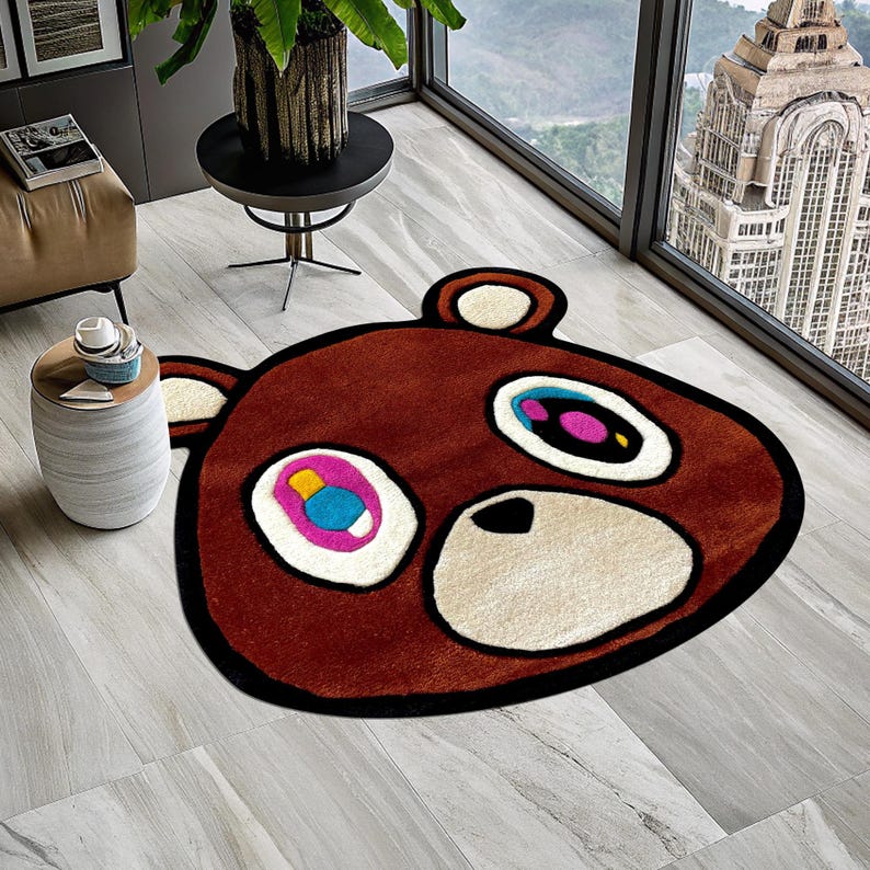 May include: A brown bear rug with a white outline and colorful eyes. The rug is made of soft, plush material and features a cartoon-like design.