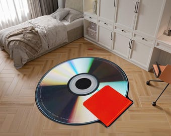 Könnte beinhalten: Ein runder, silberfarbener CD-Teppich mit einem roten quadratischen Aufkleber auf der rechten oberen Seite. Der Teppich liegt auf einem Holzboden.