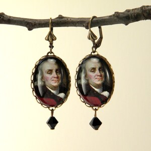 May include: A pair of antique brass-toned earrings. Each earring features an oval portrait of Benjamin Franklin, framed with a scalloped edge. A black, faceted bead dangles from the bottom of each earring. The earrings are suspended from a decorative lever-back clasp.