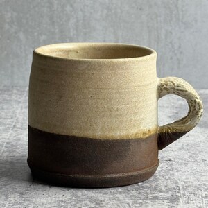 May include: A two-tone ceramic mug with a light beige upper section and a dark brown lower section. The handle has a textured, organic design. This handmade mug is suitable for hot drinks such as tea or coffee. The mug is approximately 10 cm tall.