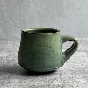 May include: A green ceramic mug with a rounded body and a handle. The mug has a textured surface and a dark rim, suggesting a rustic aesthetic. The mug is designed for hot drinks. The mug is likely handmade.