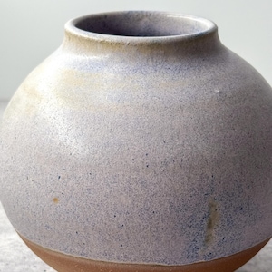 Może przedstawiać: Okrągły, ceramiczny wazon z nakrapianym, jasnoniebiesko-szarym zewnętrzem. Wazon ma szeroki, zaokrąglony korpus i wąską szyjkę. Podstawa jest ciepła, ziemista brązowa. Wazon jest przedmiotem dekoracyjnym.