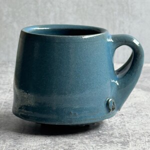 May include: A teal ceramic mug with a rounded body and a handle. The mug has an irregular shape, giving it a unique, artisanal feel. The interior is dark, and the exterior has a textured, matte finish. The mug is suitable for hot or cold drinks.