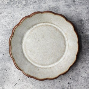 May include: A round ceramic plate with a scalloped edge, featuring a speckled off-white center glaze and a contrasting dark brown rim. The surface of the plate has a textured, matte appearance with visible clay flecks throughout the glaze.