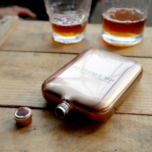 Könnte beinhalten: Eine roségoldene Metall-Taschenflasche mit der Aufschrift "Bottoms Up" auf der Vorderseite. Die Flasche ist offen und der Verschluss liegt daneben.