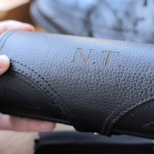 Könnte beinhalten: Ein schwarzes Lederetui mit den Initialen "N.T." auf der Oberfläche. Das Etui hat eine strukturierte Oberfläche und ein Nahtdesign entlang der Kanten. Das Etui ist zylindrisch und scheint für Brillen oder Sonnenbrillen zu sein.