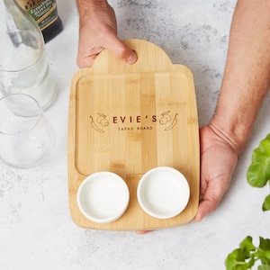 Peut inclure: Un plateau à tapas en bois avec deux petits bols blancs et le texte "Evie's Tapas Board" gravé sur la surface. Le plateau a une poignée et est fait de bambou brun clair.