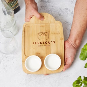 Könnte beinhalten: Ein personalisiertes Bambus-Sushi-Brett mit zwei kleinen weißen Schalen und dem eingravierten Text "JESSICA'S SUSHI BOARD" auf der Oberfläche.