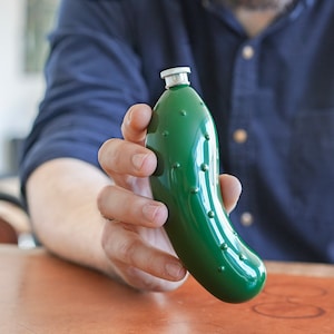 Peut inclure: Une bouteille en verre vert en forme de cornichon avec un couvercle argenté. La bouteille est tenue par la main d'une personne.