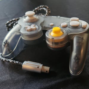 May include: A transparent gray video game controller with white buttons and a yellow joystick. The controller has a black and white braided cable and a silver connector. The controller is designed for gaming.