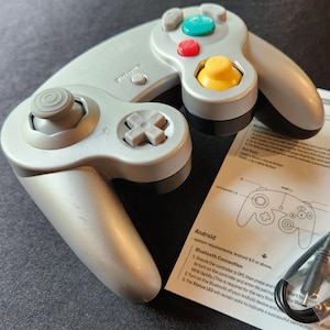 May include: A silver-gray game controller with colored buttons and a directional pad. A paper with a diagram of the controller and a USB cable are also present. The controller has a retro design.