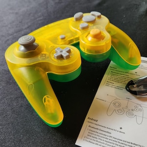 May include: A transparent yellow and green game controller with gray buttons and an orange center button. A USB cable and instruction manual are next to the controller. The manual includes the word "Android" and a diagram of the controller.