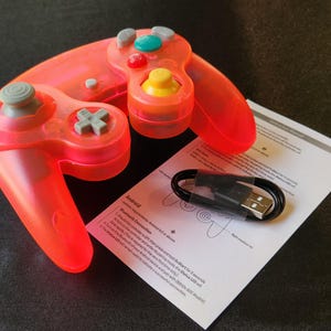 May include: A translucent orange video game controller with gray, yellow, red, and teal buttons. A USB cable and instruction manual are also present. The controller has a classic design with a directional pad and analog stick.