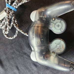 May include: A transparent gray video game controller with a braided black and white cable and a blue rubber band. The controller has two handles and several buttons. The controller is designed for gaming.