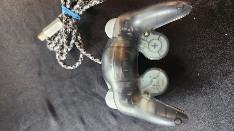 May include: A transparent gray video game controller with a braided black and white cable and a blue rubber band. The controller has two handles and several buttons. The controller is designed for gaming.