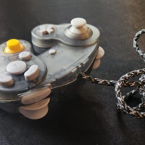 May include: A clear and gray video game controller with white and yellow buttons. The controller has a patterned cord attached, secured with a blue rubber band. The controller is designed for gaming.