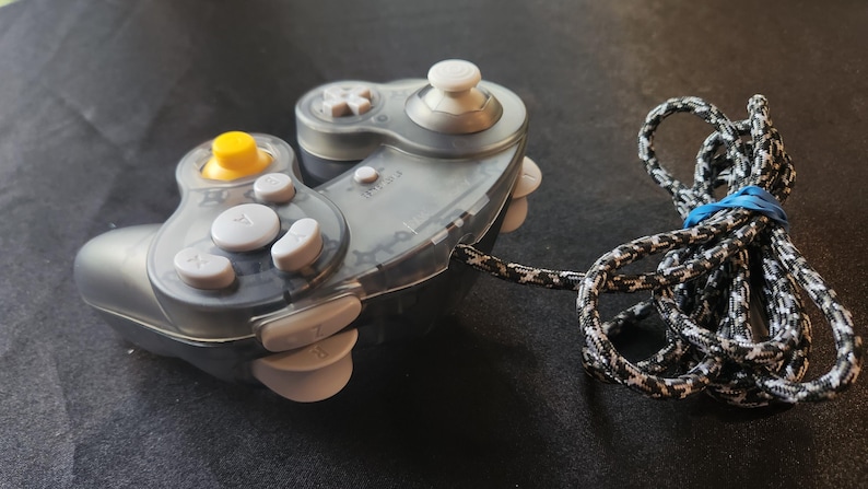May include: A clear and gray video game controller with white and yellow buttons. The controller has a patterned cord attached, secured with a blue rubber band. The controller is designed for gaming.
