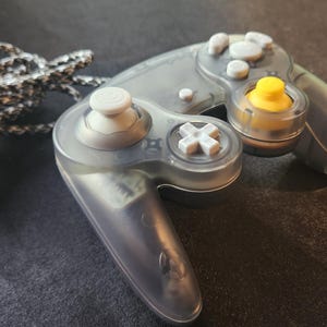 May include: A transparent gray video game controller with white and yellow buttons. The controller has a black and white braided cord secured with a blue rubber band. The controller is designed for gaming.