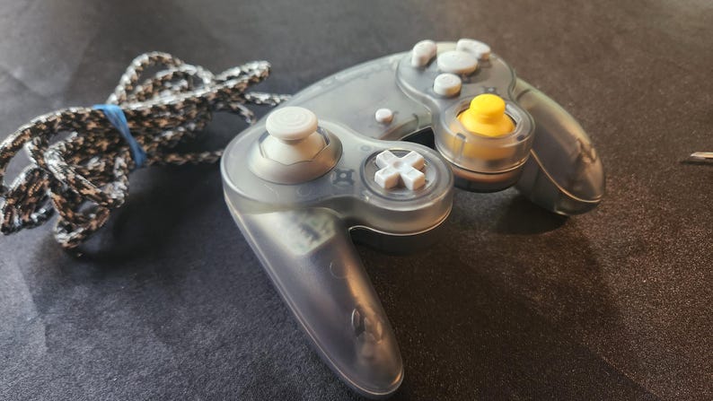 May include: A transparent gray video game controller with white and yellow buttons. The controller has a black and white braided cord secured with a blue rubber band. The controller is designed for gaming.