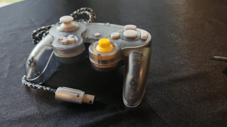 May include: A transparent gray video game controller with white buttons and a yellow joystick. The controller has a black and white braided cable and a silver connector. The controller is designed for gaming.
