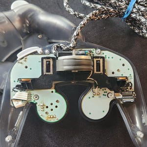 May include: A partially disassembled clear GameCube controller, revealing internal components. The controller's buttons and analog sticks are visible, along with the circuit board and wiring. A black and white braided cable is attached.