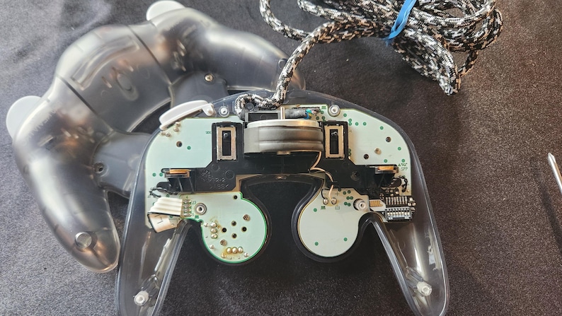May include: A partially disassembled clear GameCube controller, revealing internal components. The controller's buttons and analog sticks are visible, along with the circuit board and wiring. A black and white braided cable is attached.