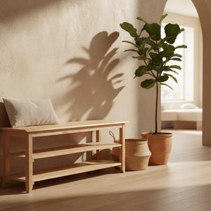 Puede incluir: Un banco de madera clara con dos estantes y un cojín beige. Una planta en maceta y una cesta de mimbre están cerca. La escena está bañada por la luz cálida del sol, con sombras en la pared.