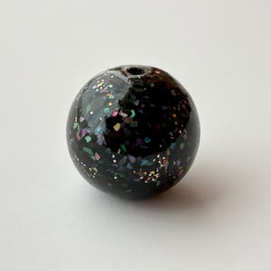 以下が含まれることがあります： 黒色の球形のビーズで、虹色のグリッターが埋め込まれています。ビーズは光沢のある仕上げで、上部に小さな穴があり、紐を通すためのものです。この装飾的なビーズは、ジュエリー作りやその他のクラフトプロジェクトに使用できます。