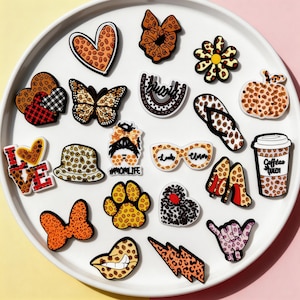 May include: A collection of decorative shoe charms in various shapes, including hearts, butterflies, and flowers. The charms feature leopard print, floral, and graphic designs. Some charms have text, such as "LOVE" and "Coffee Queen."