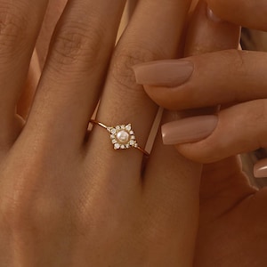Puede incluir: Un anillo de oro con una perla central rodeada por un cuadrado de pequeños diamantes. El anillo está engastado en una delicada banda de oro. El anillo se muestra en una mano con uñas largas y pulidas.