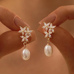 Peut inclure: Une paire de boucles d'oreilles dorées avec un motif floral orné de cristaux transparents et une perle blanche pendante. Les boucles d'oreilles sont présentées sur un fond doux.