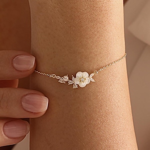 May include: A gold bracelet with a floral design, featuring white petals and sparkling accents. The bracelet is worn on a wrist, highlighting its intricate details and elegant style. The bracelet is a piece of jewellery.
