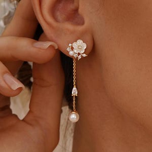 May include: A gold-tone drop earring featuring a white flower-shaped stud with small clear crystals and a delicate chain. The chain suspends a pear-shaped clear crystal and a round white pearl at the bottom, creating a layered, dangling design.