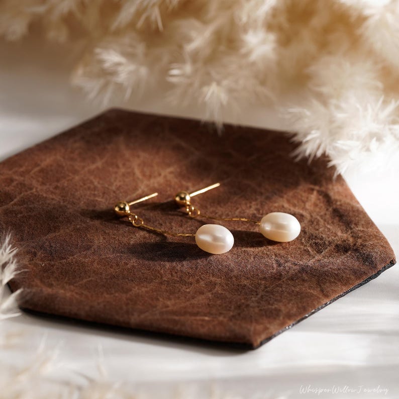 May include: A pair of gold dangle earrings with white pearl drops. The earrings are on a brown leather surface.