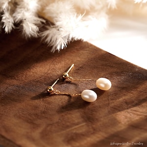 May include: A pair of gold dangle earrings with a single white pearl on each earring. The earrings are on a wooden surface.