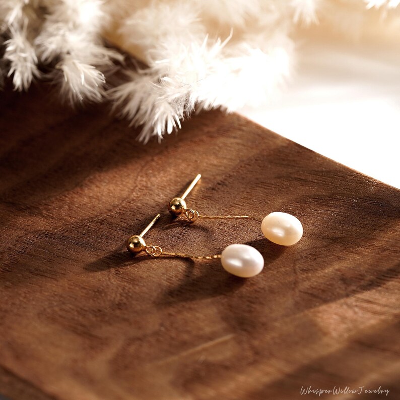 May include: A pair of gold dangle earrings with a single white pearl on each earring. The earrings are on a wooden surface.