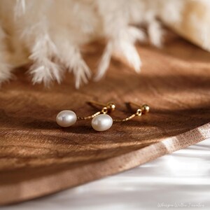 May include: A pair of gold pearl earrings with a delicate chain connecting the two pearls. The pearls are white and round, and the earrings are on a wooden surface.