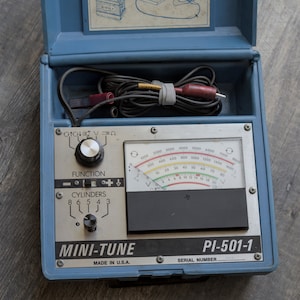 May include: A blue and silver automotive engine tune-up tool with a dial for selecting the number of cylinders and a dial for selecting the function. The tool has a meter with a needle and a scale that reads from 0 to 1000. The tool is labeled "MINI-TUNE" and "PI-501-1".