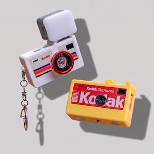 May include: A white Kodak camera keychain with a red lens ring and a square light attachment, alongside a yellow Kodak Charmera camera with a red and white logo graphic on the front. Both items feature a rectangular body and a central lens.