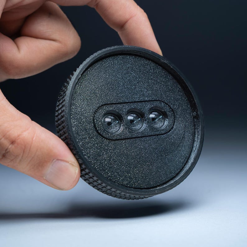 May include: A black, circular object with a textured edge and a recessed oval containing three small, clear, spherical elements. The object is held in a person's hand against a dark background.