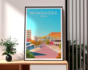Puede incluir: Lámina artística enmarcada que presenta una ilustración estilizada de Windhoek, Namibia. La obra muestra una carretera rosa que atraviesa una ciudad con edificios y una cordillera bajo un cielo azul. La palabra "Windhoek" está en la parte superior.