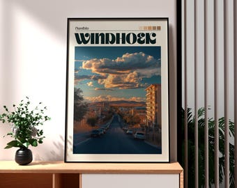 Puede incluir: Impresión artística enmarcada que representa una escena callejera en Windhoek, Namibia, bajo un cielo dramático. La obra de arte tiene un marco negro e incluye el nombre de la ciudad en letras negritas y estilizadas. La impresión se exhibe sobre una superficie de madera.