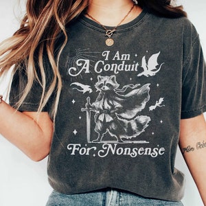 May include: A dark gray t-shirt with a white graphic of a raccoon wearing a cloak and holding a sword. The text "I Am A Conduit For Nonsense" is printed on the shirt.