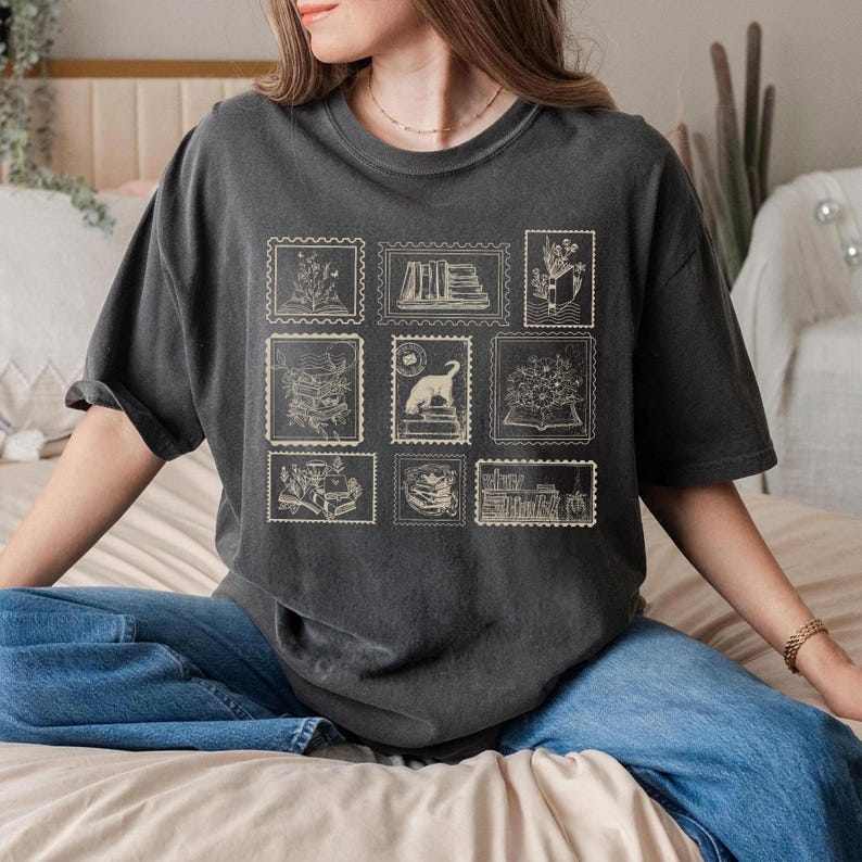 Puede incluir: Camiseta gris oscuro con ilustraciones estilo sello postal. Los sellos representan libros, flores y un gato. La camiseta tiene un corte holgado y mangas cortas.