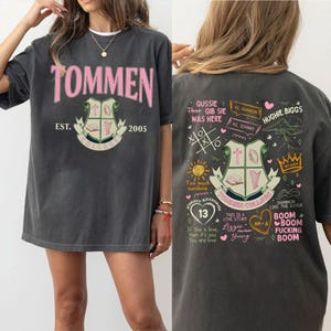 May include: A gray t-shirt with a pink graphic print of the text "TOMMEN COLLEGE" and the text "EST. 2005". The back of the shirt has a graphic print of a crest with a cross, a book, and a harp. The text "TOMMEN COLLEGE" is printed below the crest. The shirt also has other graphic prints, including a sun, a heart, and the text "THIS IS A LOVE STORY".