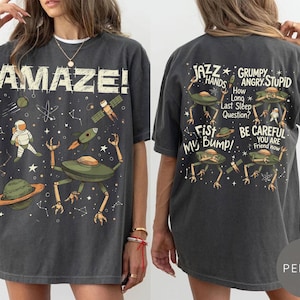 May include: Dark gray t-shirt with a space-themed design. The front features the word "AMAZE!" and illustrations of an astronaut, planets, rockets, and a space alien. The back has alien illustrations and phrases like "Jazz Hands" and "Be Careful."