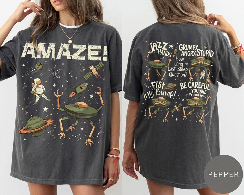 May include: Dark gray t-shirt with a space-themed design. The front features the word "AMAZE!" and illustrations of an astronaut, planets, rockets, and a space alien. The back has alien illustrations and phrases like "Jazz Hands" and "Be Careful."
